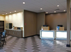Hotel lobby with a check-in counter on the right, two computers, and a sitting area on the left. The floor has a geometric pattern, and the lighting is bright.