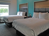 Two double beds with white bedding in a hotel room with blue accent wall and a window.