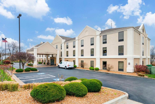 A beige three-story hotel building with a few cars parked in front, surrounded by trimmed bushes, under a partly cloudy sky.