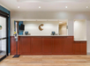 Hotel reception area with a wooden counter, two check-in monitors, and a hand sanitizer dispenser. Wall features a stylized letter "C.