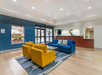 Hotel lobby with blue sofa, four yellow armchairs, and a coffee table on a patterned rug. Reception desk at the back and large windows on the left. Modern, well-lit interior.