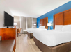 Hotel room with two double beds, wooden headboards, a flat-screen TV, a desk, and a chair. Neutral-colored walls, blue accent wall, and large window with curtains.