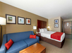 Hotel room with a double bed, blue sofa, wooden coffee table, two bedside lamps, and framed pictures on the wall. Brown accent wall behind the bed. Door visible in the background.