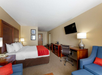 Hotel room with a king-size bed, red accents, a desk, chair, flat-screen TV, and two blue armchairs. Lamps provide ambient lighting.