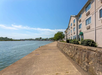 A waterside walkway beside a multi-story building on a clear day, with the river on the left and greenery in the distance.