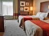 Hotel room with two double beds, red and beige bedding, a small desk and chair, three framed artworks on the wall, a tall lamp, dresser, and a window with curtains.