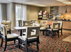 A hotel dining area with empty tables and chairs, carpeted floor, and a counter with coffee machines and breakfast items.