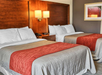 Two neatly made double beds with brown headboards, white pillows, and orange throws in a hotel room. A wall lamp and framed artwork are visible above a bedside table with a telephone.
