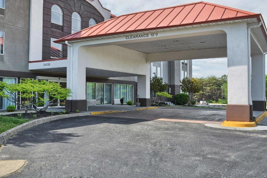 Hotel entrance with a red roofed canopy providing 12-foot clearance. The building has large windows, green landscaping, and an asphalt driveway.