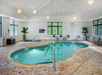 Indoor swimming pool with a kidney-shaped design, surrounded by tile flooring. There are several chairs and potted plants along the walls, with windows letting in natural light.