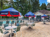 Outdoor seating area with tables and chairs under blue umbrellas near a fenced pool, surrounded by trees and a building.