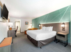 Modern hotel room with a king-sized bed, green accent wall, wall-mounted TV, desk, and chair. A small sofa and coffee table are placed in the corner. Neutral tones dominate the decor.