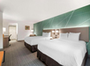 Hotel room interior with two double beds, white bedding, modern headboard, patterned green wall, desk with chair, and wall art. Neutral carpet covers the floor, and the entrance is visible.