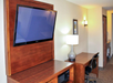 Hotel room with a wall-mounted TV, wooden desk, lamp, office chair, and an open door leading to another section of the room.