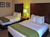 A hotel room with two double beds, each with white linens and green bed runners. A wooden nightstand with a lamp is positioned between the beds. Two framed pictures hang on the wall.