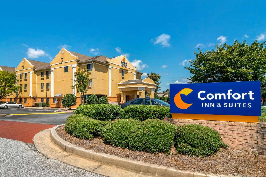 Comfort Inn & Suites building with a yellow exterior is surrounded by trees and bushes. A blue sign with the hotel logo is visible in the foreground. Clear blue sky above.