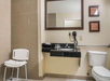 Accessible bathroom with a shower chair, sink with toiletries, wall-mounted hairdryer, and toilet. Beige tiles and safety handrail are visible.