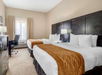 Hotel room with two double beds, white linens, brown bed runners, a desk with a chair, and a window with curtains. Dark wood furniture and neutral-colored walls.