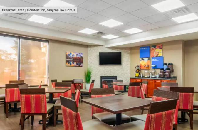 Hotel breakfast area with red-cushioned chairs and wooden tables. A TV is mounted on the wall near a corner with coffee machines and a small plant decoration.