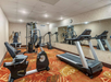 Gym room with exercise equipment, including a treadmill, stationary bike, and weight machines. The carpet is patterned, and a large mirror covers one wall.