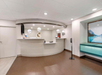 Hotel lobby with a curved reception desk, overhead lighting, and a wall-mounted TV. A large framed landscape and floor sanitizer stand are on the right.