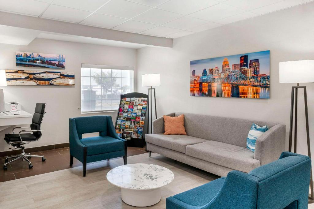Modern waiting room with a gray sofa, two blue chairs, a white round table, a desk with a chair, and cityscape artwork on the walls. A brochure stand is near the window.