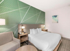 Hotel room with a double bed, green abstract wall art, nightstands with lamps, a beige armchair, and a small round table.