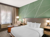 Hotel room with a double bed, an armchair, a side table, and an air conditioning unit. The wall features a green geometric pattern, and a window with curtains is on the left.