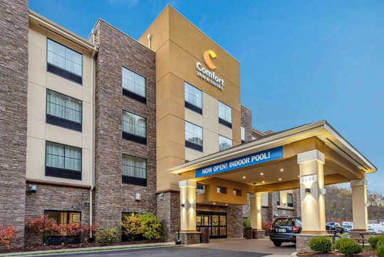 Exterior view of a Comfort Inn hotel with a sign announcing the indoor pool is now open. A car is parked under the entrance canopy.