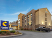 Comfort Inn & Suites building with a parking lot and a visible sign. The hotel has four stories with exterior brick and tan siding, set against a clear blue sky.