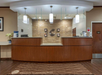Hotel reception desk with wooden finish, decorative wall, and hanging lights. A welcome mat reads "Welcome to Pittsburgh." An AED device is mounted on the wall to the right.