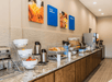 A breakfast bar with different cereals, pastries, coffee, fruit, juices, and silver serving platters with art on the wall above it and dark brown cabinets underneath.