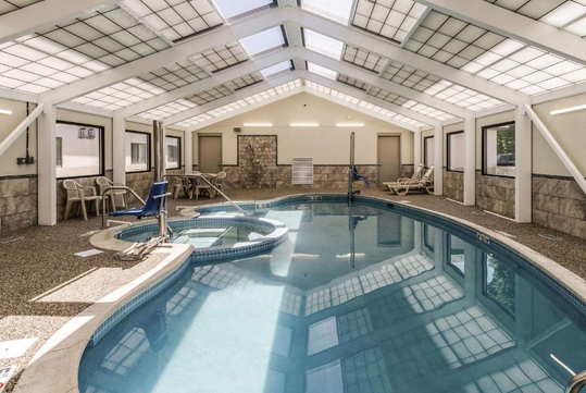 Indoor swimming pool area with a hot tub on the left, surrounded by white beams and a glass ceiling. Four loungers and three chairs are along the perimeter.