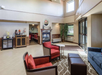 Hotel lobby with modern furniture including red and blue chairs, a round table, and a fireplace. Beverage station and dining area visible in the background. Large windows provide natural light.