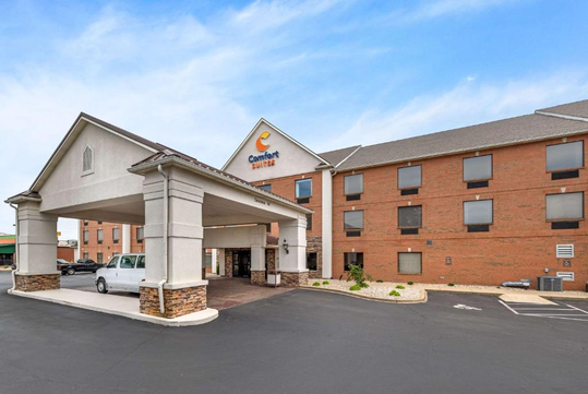A brick hotel building with a covered entrance and a white van parked nearby. The sign reads "Comfort Suites.