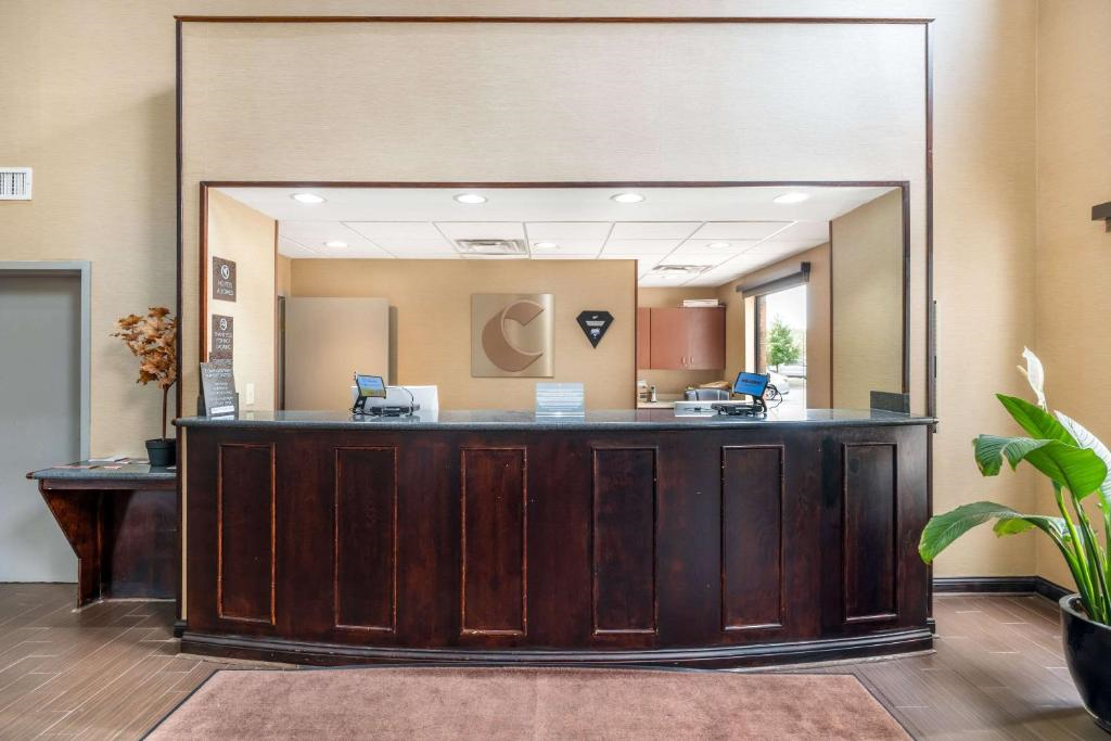 Hotel reception desk with dark wood finish, two computers, wall art, and a potted plant on the right. A door is on the left, and a window provides natural light in the background.