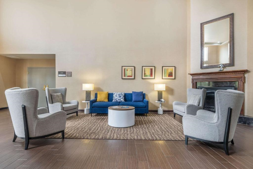 A hotel lobby with four gray armchairs and a blue sofa arranged around a round table on a rug. Three framed artworks and a mirror are on the walls. A fireplace is visible on the right.