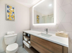 Modern bathroom with a large illuminated mirror above a wooden vanity, sink, and open shelving with towels. A toilet is on the left, and a geometric framed artwork hangs on the wall.