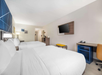 Hotel room with two double beds, white linens, a wall-mounted TV, desk with a chair, and a small bench. Neutral-colored walls and natural light from a window offer a simple, modern ambiance.