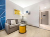 Modern hotel room with a gray sofa, yellow coffee table, floor lamp, and decorative wall art. Door at the end of a short hallway. Bright and clean space.