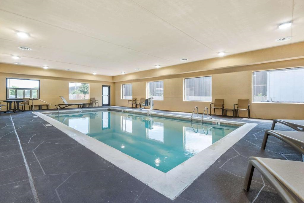 Indoor swimming pool with lounge chairs, tables, and large windows providing natural light. The pool is surrounded by a smooth, dark flooring and beige walls.