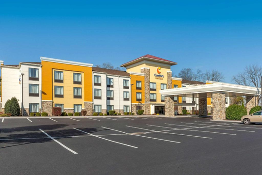 A four-story hotel with yellow and white exterior, a covered entrance, and an empty parking lot under a clear blue sky.