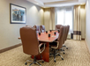A conference room with a long wooden table, eight brown leather chairs, coffee mugs, and notepads. A framed picture is on the wall, and a window is at the end of the room.