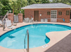 Outdoor swimming pool with a brick building and chairs in the background. The pool area is surrounded by a patio with safety signage visible.