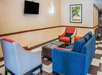 Hotel lobby with a blue sofa, two chairs, an ottoman, and a wall-mounted TV. A framed picture of a tree-lined path hangs on the wall.