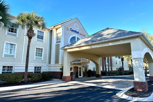 Front exterior view of a Comfort Suites hotel, featuring a covered entrance and surrounding greenery under a clear blue sky.