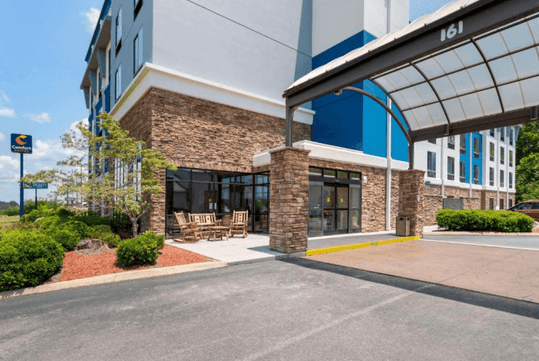 Hotel entrance with stone facade, glass doors, and a covered driveway. Wooden benches are placed along the walkway. A sign with a hotel logo is visible in the background.