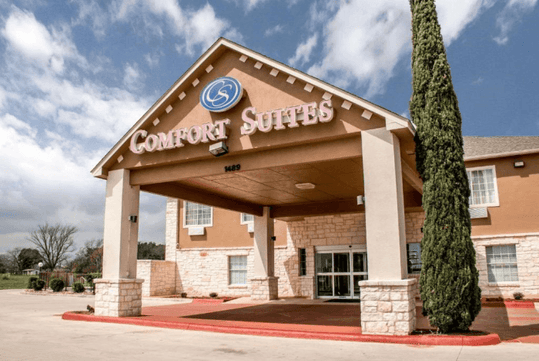 The front entrance of the Comfort Suites New Braunfels on a bright sunny day with clouds in the sky.