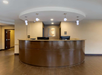 A hotel reception area with a curved wooden desk, two computer monitors, overhead pendant lights, and a wall with a logo behind it.