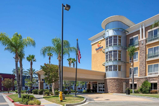 A hotel exterior with a modern design, palm trees, and an American flag. The building has multiple floors and features a circular glass entrance.